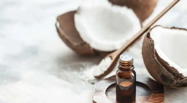 A small bottle of coconut oil sits near a halved coconut and a wooden spoon with coconut flakes. This image prompts the question: is coconut oil safe for cats?