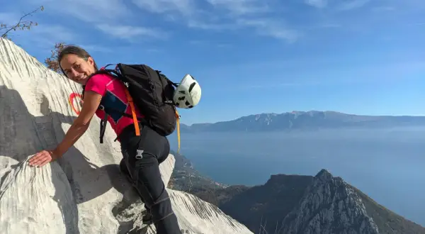 Donna sorridente su roccia con zaino e casco, vista lago e montagne. Un facile sentiero sul monte Pizzocolo.
