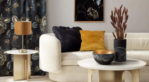 Elegancki salon z kanapą, ozdobnymi poduszkami i wazonem z suszonymi trawami zamiast kwiatów. Złote akcenty i ciemne zasłony tworzą przytulną atmosferę.