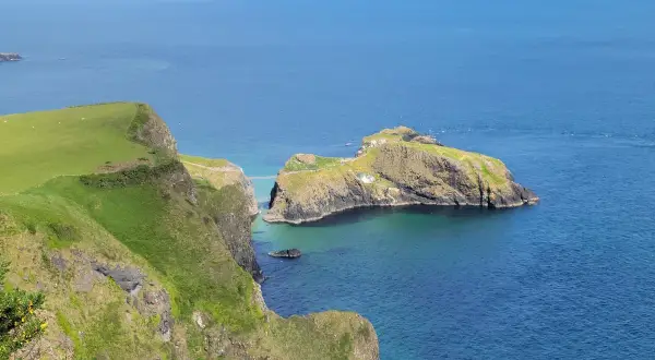 Falaises verdoyantes de la Causeway Coast, avec une île rocheuse reliée par un pont suspendu. L'eau bleue claire invite à la découverte.
