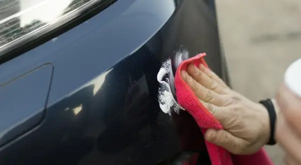 Ręka z czerwoną ściereczką poleruje ciemny lakier samochodu, usuwając drobne rysy. Rozważając powłoka ceramiczna wady i zalety, warto pamiętać o takich zabiegach.