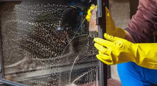 Mit gelben Handschuhen wird das Kaminglas gereinigt. Schaum und Schlieren bedecken die Scheibe, während eine Hand mit einem Schwamm schrubbt.