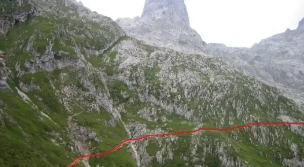 Sendero marcado en rojo asciende por la ladera verde hacia el imponente Sotres Urriellu, una cumbre rocosa en los Picos de Europa.