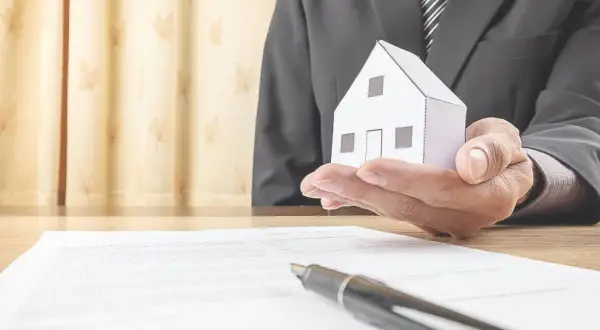 Un uomo in giacca e cravatta tiene una casetta di carta, simbolo di un accordo di leasing immobiliare per privati.