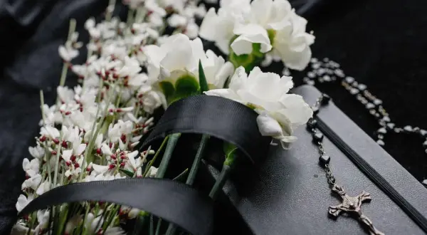 Białe kwiaty i drobne gałązki, przewiązane czarną wstęgą, symbolizują smutek i pamięć. Obok leży różaniec i notes, podkreślając powagę chwili. To piękna florystyka funeralna.