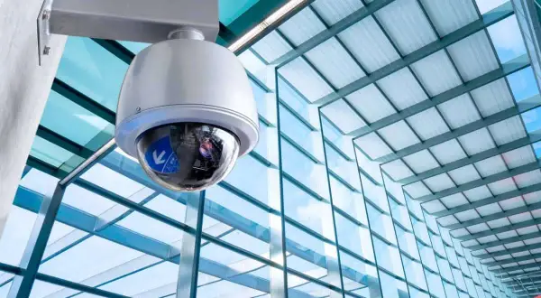 Caméra de surveillance fixée à un mur, prête à filmer. Le bâtiment moderne avec sa façade vitrée reflète le ciel bleu.
