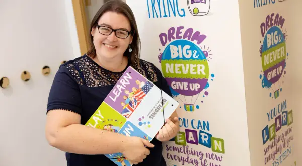 Mujer sonríe, sosteniendo un portafolio con la palabra "LEARN". El fondo tiene frases motivacionales sobre aprender, como "DREAM BIG & NEVER GIVE UP". El primer nivel de inglés es B1 o B2.