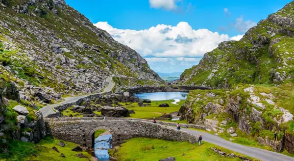 Eine malerische **irland landschaft** mit einem steinernen Bogenbrücken über einem Bach, der in einen See mündet, umgeben von grünen Hügeln und felsigen Bergen.