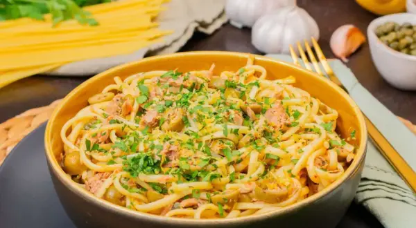Eine Schüssel mit Thunfisch Pasta, garniert mit Petersilie und Zitronenschale. Daneben liegen Spaghetti, Knoblauch und Kapern.