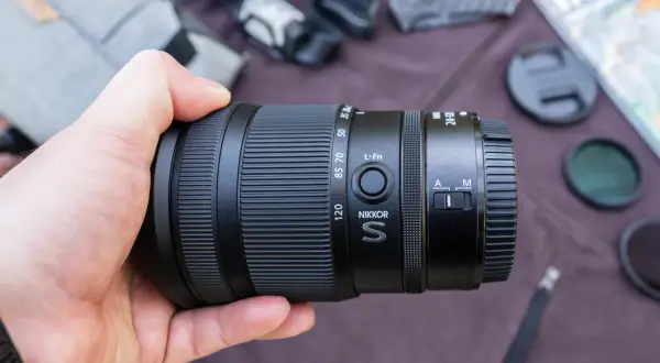 Dłoń trzyma obiektyw Nikkor S 24-120mm, idealny do pełnej klatki Nikon. W tle akcesoria fotograficzne.