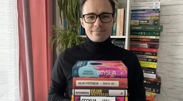 Mężczyzna w okularach trzyma stos książek, wśród nich "Odyseja" i dzieła Colsona Whiteheada. W tle regały pełne literatury, przypominające o bogactwie polskiej i światowej prozy, jak u Bolesława Prusa.