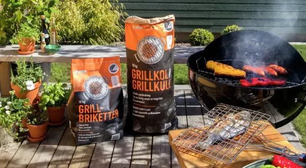 Två säckar med grillkol eller briketter står redo för grillkvällen. Majskolvar, korvar och paprika ryker på grillen.