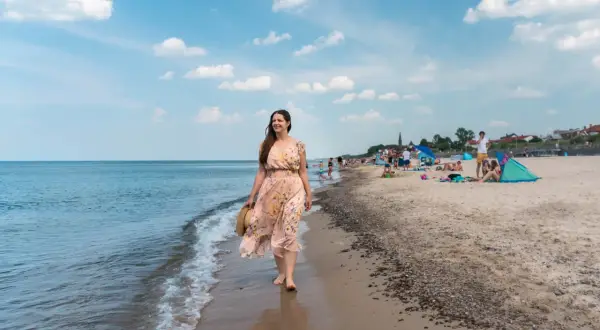 Kobieta w kwiecistej sukience spaceruje brzegiem morza. W tle plaża pełna ludzi, parawanów i namiotów – typowe sarbinowo atrakcje.