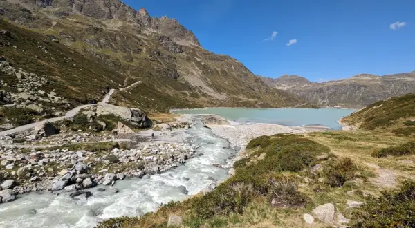 Berglandschaft mit türkisfarbenem Wasser des Silvretta Stausees, der von einem Fluss gespeist wird. Wanderweg schlängelt sich den Hang hinauf.