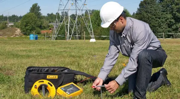 Technicien en plein air vérifiant la valeur prise de terre d'une maison, près d'un pylône électrique.