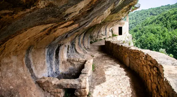 Sentiero roccioso conduce alla chiesetta di San Bartolomeo in Legio, incastonata nella roccia con vista su boschi rigogliosi.