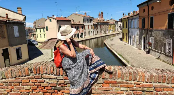 Donna con cappello di paglia e zaino rosso seduta su un ponte di mattoni a Comacchio, con canali e case colorate. Un luogo ideale per una gita con bambini.