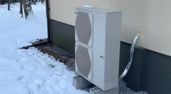 Jednostka zewnętrzna pompy ciepła stoi na betonowych podstawach w zasypanym śniegiem ogrodzie. Czy pompa ciepła jest głośna? Zimą, gdy wszystko wokół jest ciche, można to sprawdzić.