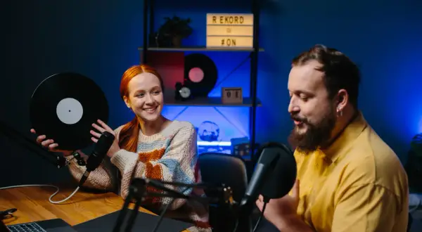 Coppia discute di come fare un podcast, lei tiene un vinile, lui parla al microfono.