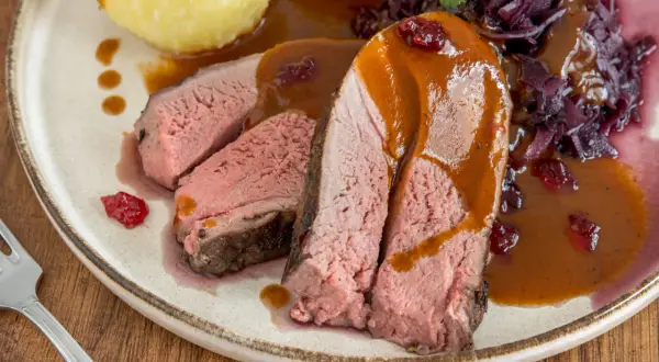 Saftiges Rehschulter Rezept: Rosa gebratenes Fleisch mit Sauce, Rotkohl und Klößen auf einem Teller.