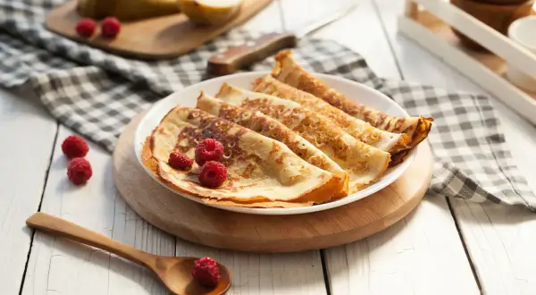Frisch zubereitete Pfannkuchen, ein köstliches Pancakes Rezept, garniert mit Himbeeren auf einem Teller. Daneben liegen Birnen und ein Holzlöffel.