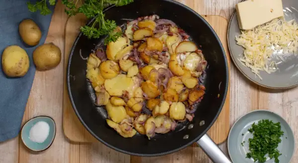 Bratkartoffeln aus rohen Kartoffeln, Zwiebeln und Speck in der Pfanne, dazu geriebener Käse und Petersilie.