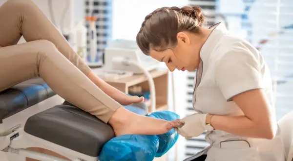 Profesjonalny pedicure co to? Kosmetyczka w białej bluzce i rękawiczkach dba o stopy klientki na wygodnym fotelu.
