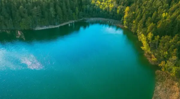 Głębokie, turkusowe jezioro otoczone gęstym lasem. Woda lśni w słońcu, tworząc odbicia chmur.