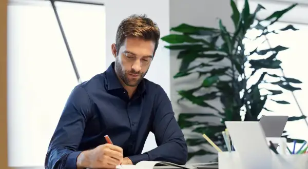 Hombre concentrado escribiendo en un diario en su escritorio, rodeado de plantas y un portátil.