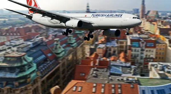 Samolot Turkish Airlines przelatuje nad miastem, jakby zmierzał w kierunku **Zakaukazia**. W tle widać zabytkowe budynki z czerwonymi dachami.