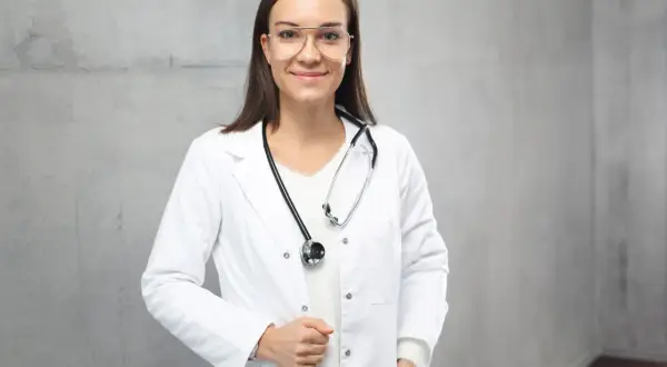 Lekarka w białym fartuchu ze stetoskopem na szyi, na tle szarej ściany.
