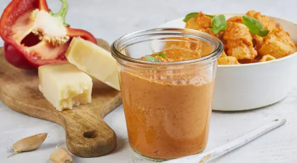 Ein Glas mit cremigem Paprika Pesto, daneben ein halber roter Paprika, Parmesan und Knoblauch. Im Hintergrund eine Schüssel Pasta mit dem Pesto.
