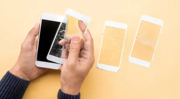 Ręce trzymają biały telefon z pękniętym szkłem hartowanym 5d. Obok leżą dwa inne telefony z rozbitymi ekranami.