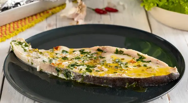 Un succulento pesce spada al forno, condito con erbe aromatiche e olio d'oliva, servito su un piatto scuro.
