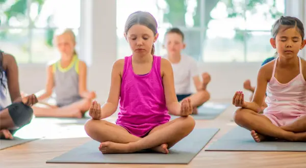 Niños practicando ejercicios de respiración y meditación en un salón de clases, sentados en colchonetas con los ojos cerrados.