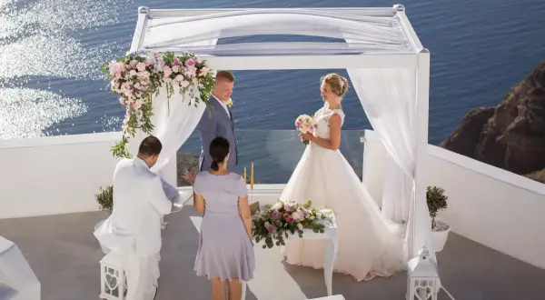 Piękny ślub w Grecji. Para młoda stoi pod białym baldachimem, w tle błękitne morze. Ceremonia odbywa się na tarasie z widokiem na ocean.