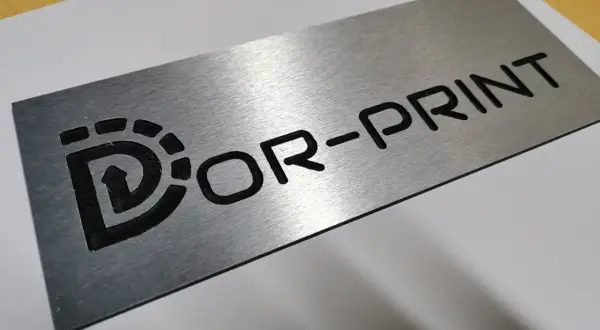 Metalowa tabliczka grawer z logo "Dor-Print". Wygląda jakby była wykonana z aluminium.