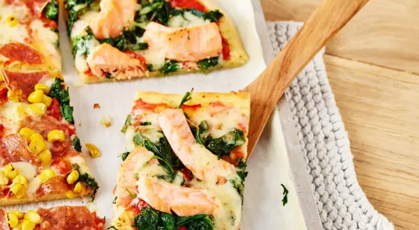 Pizza mit Lachs und Spinat, belegt mit geschmolzenem Käse, wird mit einem Holzschieber serviert.