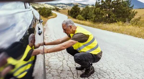 Hombre con chalecos reflectantes, agachado junto a un coche averiado en una carretera rural.