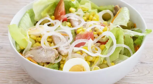 Deliciosa ensalada mixta con atún, huevo duro, maíz, tomate y cebolla, servida en un cuenco blanco sobre una mesa de madera.