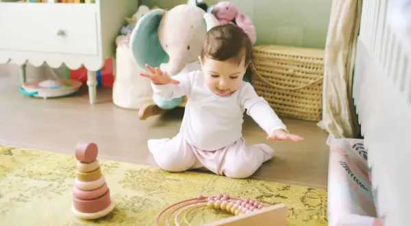 Ein **baby 9 monate** sitzt auf einem Teppich und spielt mit Holzspielzeug. Im Hintergrund sind Stofftiere und ein Kinderbett zu sehen.