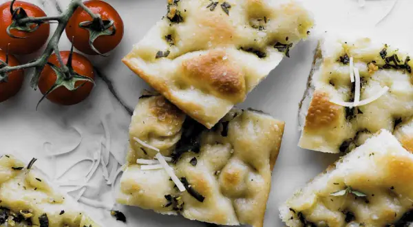 Pyszna focaccia z ziołami i parmezanem, idealna do przygotowania według prostego przepisu. Obok gałązka pomidorków koktajlowych.