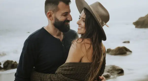 Ett par omfamnar varandra vid havet, deras blickar möts i en tydlig attraktion mellan två personer.