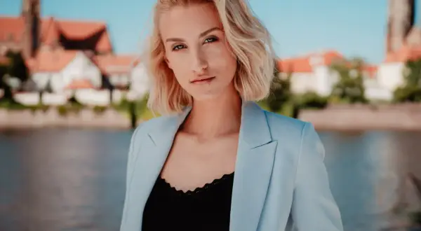 Blondynka w błękitnej marynarce pozuje w plenerze nad rzeką, z widokiem na zabytkowe budynki w tle.