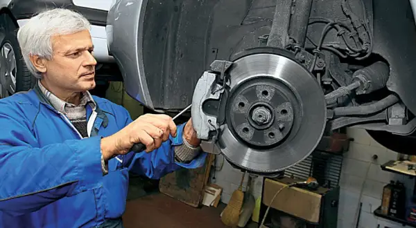 Mechanik w niebieskim kombinezonie pracuje nad tarczą hamulcową samochodu, dbając o sprawność układu hamulcowego.
