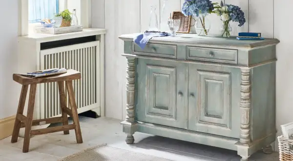 Ein blau-graues Sideboard mit abgenutzter Patina, das den "shabby chic"-Stil verkörpert. Daneben ein Holzhocker und Blumen.