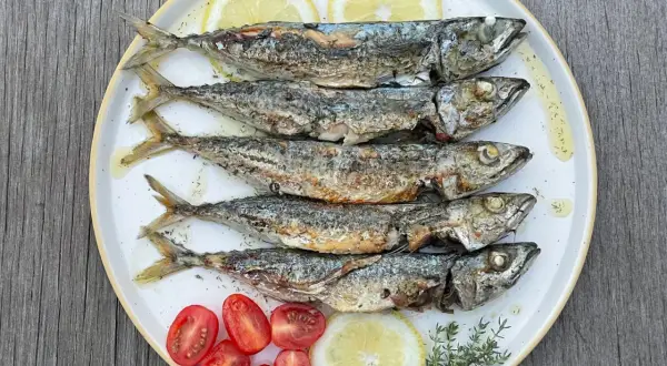 Cinq sardines au four, dorées et savoureuses, servies avec des quartiers de citron, des tomates cerises et des herbes fraîches sur une assiette blanche.