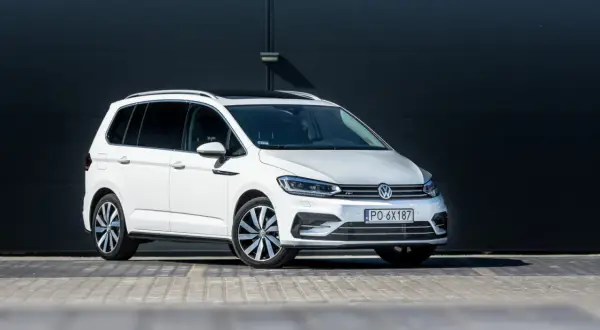 Biały Volkswagen Touran z czarnym dachem i przyciemnianymi szybami, zaparkowany przed ciemną, nowoczesną ścianą.