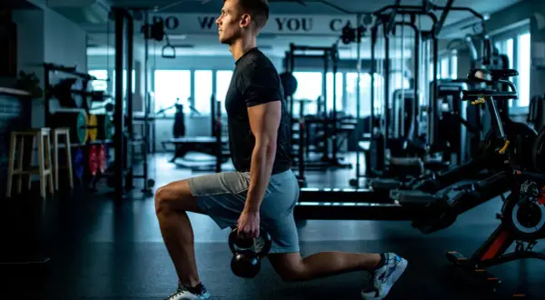 Mann macht Ausfallschritte mit Kettlebell im Fitnessstudio. Verschiedene Ausfallschritte Varianten trainieren Beine und Po.
