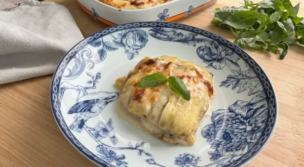 Porzione di lasagne di zucchine gratinate su piatto decorato, con basilico fresco e una teglia sullo sfondo.
